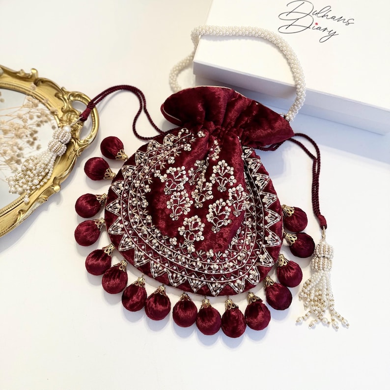 May include: A maroon velvet drawstring bag with gold and pearl embroidery and a tassel. The bag is decorated with maroon pom-poms.