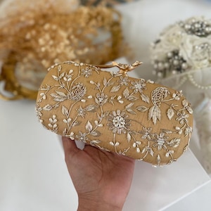 Könnte beinhalten: Eine goldfarbene, rechteckige Clutch mit aufwendigen silbernen Metallfadenstickereien in Blumen- und Blattmustern, akzentuiert durch kleine weiße Kunstperlen im gesamten Design und einen metallischen, vogelgeformten Verschluss an der Oberkante.