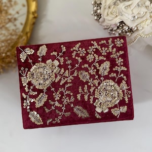 以下が含まれることがあります： 複雑な金色の刺繍とスパンコールの花柄装飾が施された、長方形のバーガンディ色のベルベットボックス。デザインには、2つの大きな中央の花のモチーフと、それを取り囲む小さな小枝、葉、繊細な蔓のようなディテールが含まれています。