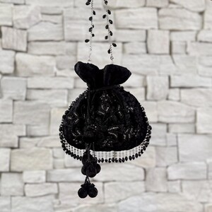 以下が含まれることがあります： 黒いベルベットの巾着バッグ。ビーズとスパンコールの複雑な装飾が施されています。バッグにはビーズのストラップとタッセルのアクセントが付いています。白いレンガの壁を背景に吊り下げられています。