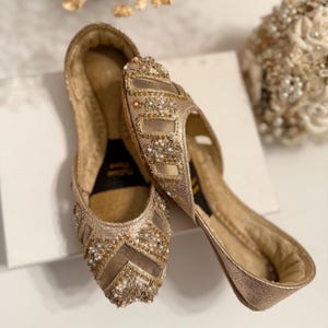 May include: A pair of gold-colored, embellished flats. The shoes feature intricate beadwork and floral designs, with a mesh overlay on the toe box. The soles are a darker gold, and the shoes appear to be unworn. The overall style is elegant and suitable for special occasions.