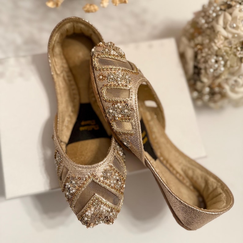 May include: A pair of gold-colored, embellished flats. The shoes feature intricate beadwork and floral designs, with a mesh overlay on the toe box. The soles are a darker gold, and the shoes appear to be unworn. The overall style is elegant and suitable for special occasions.