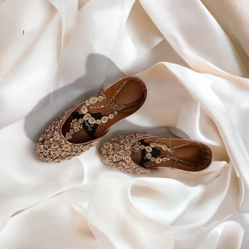 Puede incluir: Un par de bailarinas doradas adornadas con intrincados abalorios y una suela de cuero marr&oacute;n. Los zapatos est&aacute;n adornados con cristales brillantes y descansan sobre una superficie de sat&eacute;n blanco, mostrando un dise&ntilde;o lujoso.