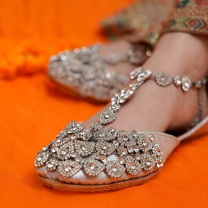 Puede incluir: Bailarinas plateadas adornadas con detalles de pedrer&iacute;a. Los zapatos presentan un dise&ntilde;o de correa en T y est&aacute;n adornados con brillantes acentos. El calzado se encuentra sobre un fondo naranja, resaltando la artesan&iacute;a ornamental.