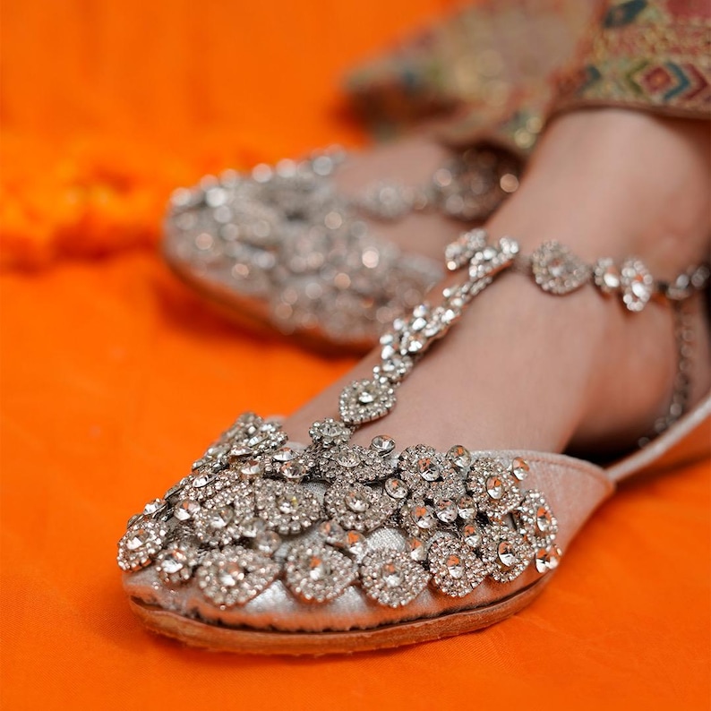 Puede incluir: Bailarinas plateadas adornadas con detalles de pedrer&iacute;a. Los zapatos presentan un dise&ntilde;o de correa en T y est&aacute;n adornados con brillantes acentos. El calzado se encuentra sobre un fondo naranja, resaltando la artesan&iacute;a ornamental.
