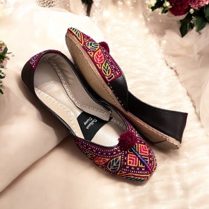 May include: A pair of black leather embroidered shoes with a maroon base and colorful floral patterns. The shoes have a pointed toe and a small pom-pom on the top. The inscription "Dilkash" is visible on the inside of one shoe.