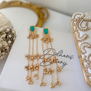 May include: Gold-coloured dangle earrings with turquoise-coloured stone accents. The earrings feature intricate detailing and dangling chains with small, round embellishments. The earrings are displayed on a white surface with the text "Dulhans Diary".