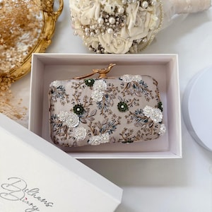 May include: A rectangular clutch bag in a light beige fabric featuring intricate embroidery of branches and leaves with white and dark green sequin floral accents. The bag has a gold-toned metal bird-shaped clasp closure on the top edge.