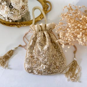May include: A gold velvet drawstring bag with intricate floral embroidery and beaded tassels. The bag features a gathered top and a small handle. The design includes gold sequins and beads, creating a luxurious look. The bag is likely for special occasions.