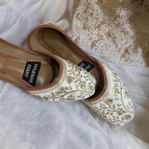 K&ouml;nnte beinhalten: Ein Paar wei&szlig;e, bestickte flache Schuhe mit goldenen F&auml;den und Perlenakzenten. Die Schuhe haben einen braunen Lederbesatz und die Worte "Dulhans Diary" sind auf der Innensohle aufgedruckt.