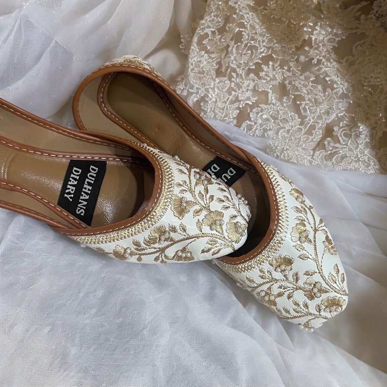 K&ouml;nnte beinhalten: Ein Paar wei&szlig;e, bestickte flache Schuhe mit goldenen F&auml;den und Perlenakzenten. Die Schuhe haben einen braunen Lederbesatz und die Worte "Dulhans Diary" sind auf der Innensohle aufgedruckt.