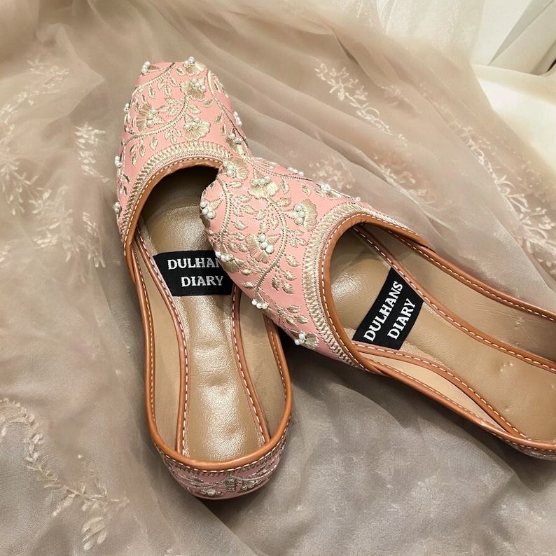K&ouml;nnte beinhalten: Ein Paar flache, rosa bestickte Schuhe mit Perlenbesatz. Die Schuhe haben eine braune Ledersohle und sind mit "Dulhan Diary" beschriftet.