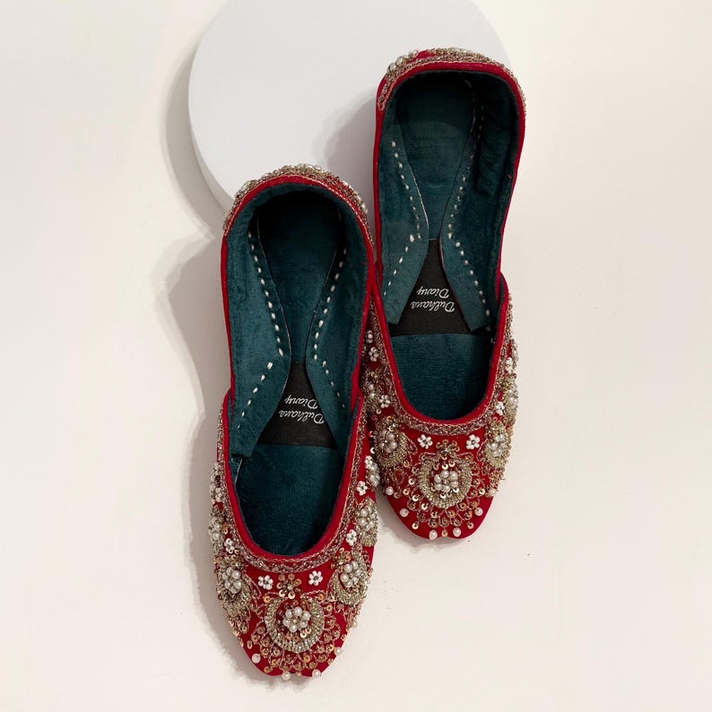 K&ouml;nnte beinhalten: Ein Paar rote Samtschuhe mit aufwendigen Silber- und Perlen-Stickereien. Die Schuhe haben eine spitze Zehenpartie und eine flache Sohle. Das Innere der Schuhe ist mit einem dunkelgr&uuml;nen Stoff gef&uuml;ttert. Der Markenname "Dulhania Dream" ist auf der Innenseite jedes Schuhs aufgedruckt.