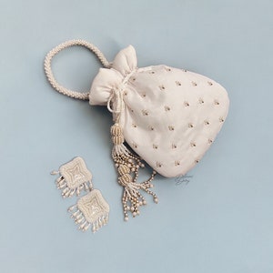 May include: A white drawstring bag with a pearl handle and tassel. The bag is embroidered with small white beads and has a pearl-embellished tassel. Two matching pearl earrings are also pictured.