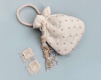 May include: A white drawstring bag with a pearl handle and tassel. The bag is embroidered with small white beads and has a pearl-embellished tassel. Two matching pearl earrings are also pictured.