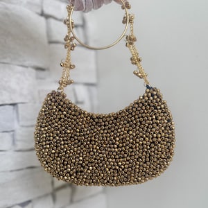 May include: A gold beaded handbag with a crescent shape. The bag features a large gold ring handle and delicate gold chain straps adorned with small gold beads. The bag is covered in small, round gold beads.