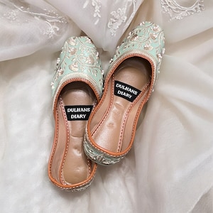 K&ouml;nnte beinhalten: Ein Paar flache, mintgr&uuml;ne bestickte Schuhe mit brauner Ledersohle. Die Schuhe haben ein silbernes und wei&szlig;es Blumenmuster und sind auf der Innensohle mit "Dulhans Diary" beschriftet.
