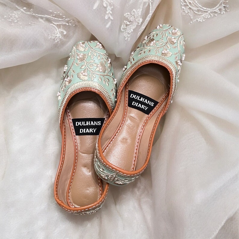 K&ouml;nnte beinhalten: Ein Paar flache, mintgr&uuml;ne bestickte Schuhe mit brauner Ledersohle. Die Schuhe haben ein silbernes und wei&szlig;es Blumenmuster und sind auf der Innensohle mit "Dulhans Diary" beschriftet.