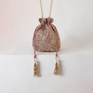May include: A dusty rose drawstring bag with gold floral embroidery. The bag has a pearl-beaded handle and tassel accents. The bag is approximately 7 inches tall and 6 inches wide.