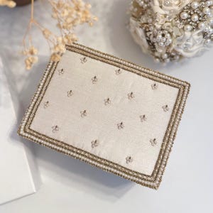 May include: Ivory rectangular clutch with a beaded border and scattered floral embellishments. The clutch features a pearl and gold beaded trim. The background includes a white surface and a bouquet of white flowers.