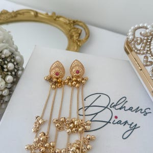 May include: Gold-toned dangle earrings with a teardrop design, featuring a red center stone and intricate detailing. The earrings have multiple strands of small gold beads. The text "Dulhans Diary" is visible on the white surface.