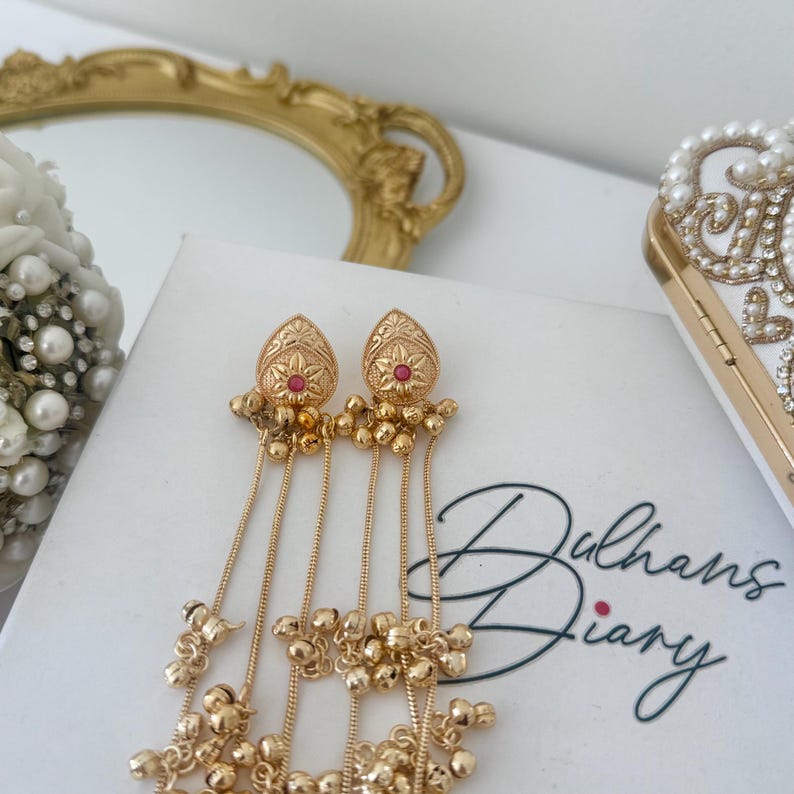 May include: Gold-toned dangle earrings with a teardrop design, featuring a red center stone and intricate detailing. The earrings have multiple strands of small gold beads. The text "Dulhans Diary" is visible on the white surface.