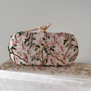 May include: An oval-shaped clutch with a floral and bird embroidery design. The clutch is beige with pink and green floral accents and a gold bird clasp. The clutch is resting on a white surface with gold embroidery.