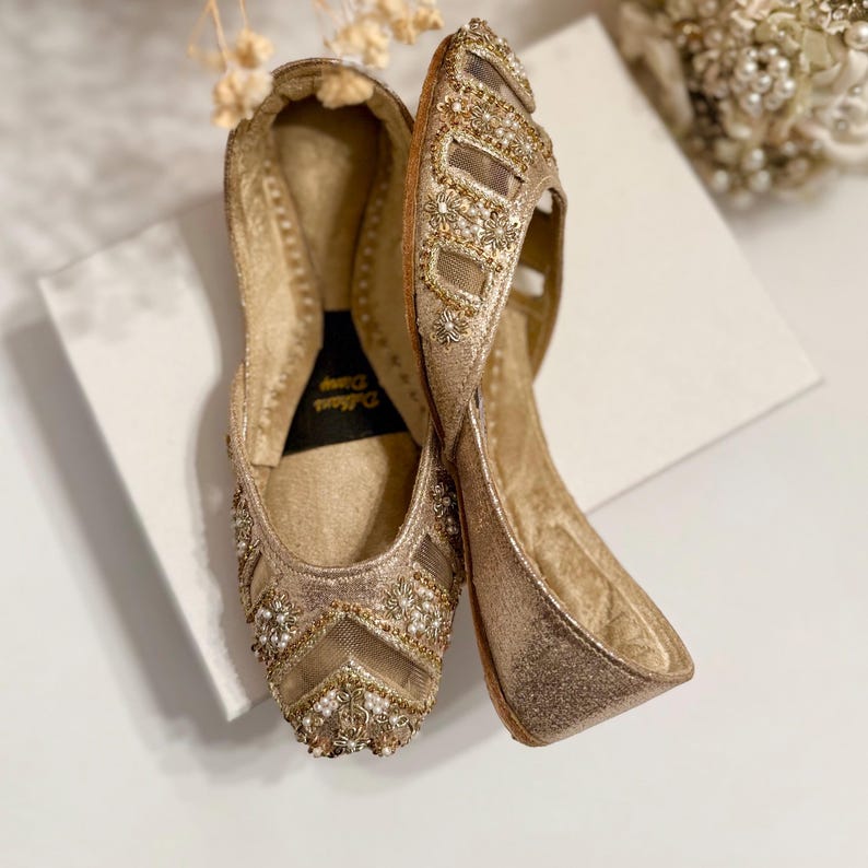 May include: A pair of gold-colored, embellished flats. The shoes feature intricate beadwork and a mesh design on the toe. The soles are a solid gold color. The shoes are displayed on a white surface.