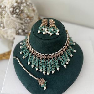 May include: An elegant jewelry set featuring a necklace, earrings, and a headpiece. The set includes turquoise-colored beads and white pearls, with gold-toned metal accents. The necklace and earrings are displayed on a dark green velvet stand.