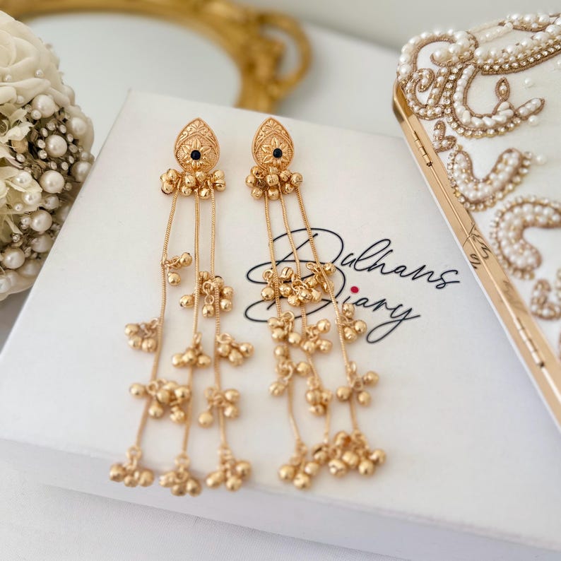 May include: A pair of gold-colored dangle earrings with intricate detailing. Each earring features a teardrop-shaped top with a small black stone and multiple strands of small gold beads. The earrings are displayed on a white box with the text "Dulhans Diary".