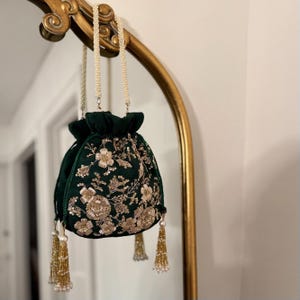 Pode incluir: Uma bolsa com cordão de veludo verde escuro com bordados florais dourados e borlas de contas. A bolsa pende de um arco decorativo dourado com uma alça de contas peroladas. O design da bolsa apresenta padrões florais intrincados.