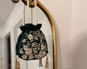 以下が含まれることがあります： 金色の花の刺繍とビーズのタッセルが付いた、濃い緑色のベルベット巾着袋。バッグは、真珠のようなビーズストラップが付いた金色の装飾的なアーチから吊り下げられています。バッグのデザインは、複雑な花のパターンが特徴です。