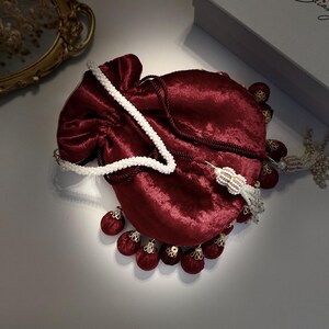May include: A burgundy velvet drawstring bag with a white pearl chain strap and gold accents. The bag has a tassel with multiple burgundy pom-poms.