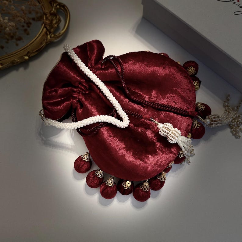 May include: A burgundy velvet drawstring bag with a white pearl chain strap and gold accents. The bag has a tassel with multiple burgundy pom-poms.