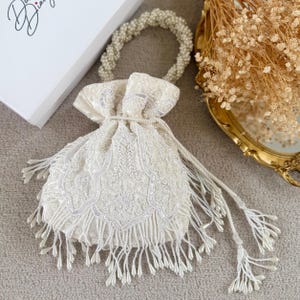 May include: Ivory drawstring bag with detailed beadwork and fringe. The bag has a beaded handle and a decorative design on the front. The bag is suitable for special occasions.