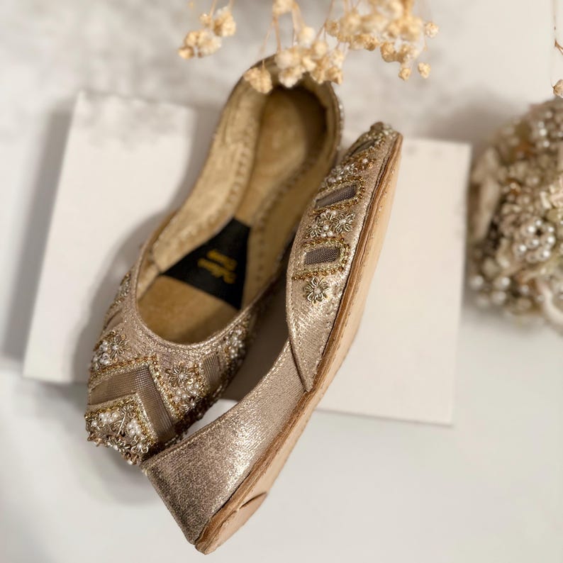 May include: A pair of gold-colored, embellished flats. The shoes feature intricate beadwork and floral accents. The design includes a mesh panel on the top of the foot. The soles are a light tan color. The shoes are displayed on a white surface.
