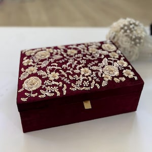 May include: A burgundy velvet box with intricate gold floral embroidery on the lid. The box has a rectangular shape with a gold clasp. The floral design includes various flower and leaf patterns.