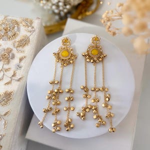 May include: A pair of gold-toned earrings with a yellow center stone and dangling chain accents. The earrings have a floral design and are displayed on a white surface. The earrings are a statement piece of jewelry.