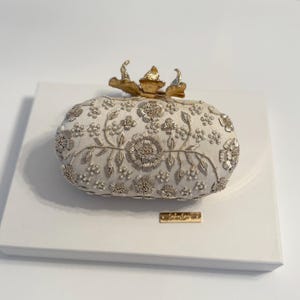 May include: A white oval clutch purse with a gold floral design and a gold flower-shaped clasp. The purse is on a white surface.