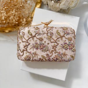 Könnte beinhalten: Eine rechteckige Clutch aus zartrosa Stoff mit aufwendigen Goldperlen, Pailletten und gestickten Blumenmustern mit Vogelmotiven. Der Verschluss ist eine goldfarbene Metallvogel-Figur, die auf der Oberkante der Tasche sitzt.