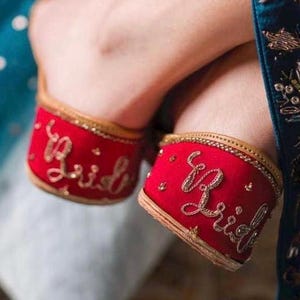 Puede incluir: Zapatos de novia rojos con bordado dorado con la palabra "Bride". Los zapatos tienen un ribete dorado y est&aacute;n decorados con peque&ntilde;os detalles dorados. Los zapatos son de estilo tradicional.