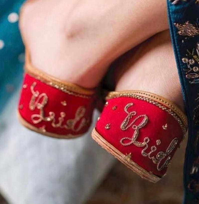 Puede incluir: Zapatos de novia rojos con bordado dorado con la palabra "Bride". Los zapatos tienen un ribete dorado y est&aacute;n decorados con peque&ntilde;os detalles dorados. Los zapatos son de estilo tradicional.