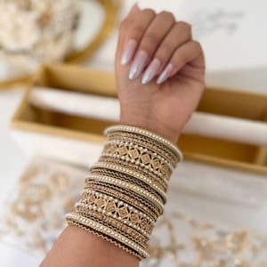 May include: A stack of gold-toned bangles with intricate geometric patterns and small white beads. The bangles are stacked on a wrist, showcasing the jewelry's design and texture. The overall aesthetic is elegant and ornate.