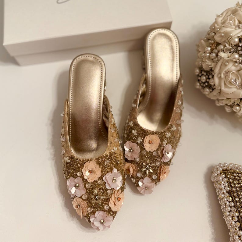 以下が含まれることがあります： ピンクとピーチの花のアクセントが付いた、ゴールドの装飾が施されたブライダルシューズのペア。シューズはポインテッドトゥで、ヒールは低くなっています。