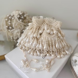May include: Ivory beaded clutch with a drawstring closure. The clutch is heavily embellished with small, round beads and larger, teardrop-shaped beads that cascade down the sides. A matching beaded tassel hangs from the drawstring. A second, similar clutch is in the background.