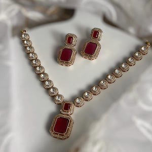 May include: A gold necklace with a red gemstone pendant and matching earrings. The necklace is made of small round stones and the pendant is a large square stone.