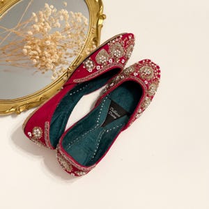 K&ouml;nnte beinhalten: Ein Paar rote Samtschuhe mit aufwendigen Gold- und Perlen-Stickereien. Die Schuhe haben eine spitze Zehenpartie und einen niedrigen Absatz. Der Markenname "Dilkash Designs" ist auf der Innensohle sichtbar.