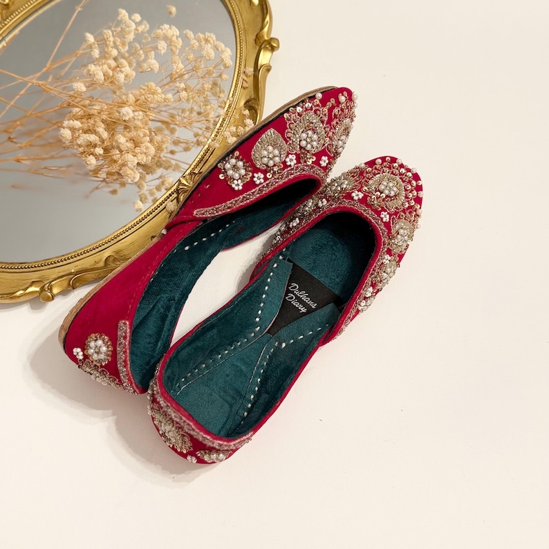K&ouml;nnte beinhalten: Ein Paar rote Samtschuhe mit aufwendigen Gold- und Perlen-Stickereien. Die Schuhe haben eine spitze Zehenpartie und einen niedrigen Absatz. Der Markenname "Dilkash Designs" ist auf der Innensohle sichtbar.