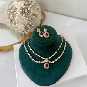 May include: A two-strand necklace and matching earrings set with red gemstones and small white stones. The jewelry is displayed on a green velvet stand. A bouquet of white flowers is in the background.