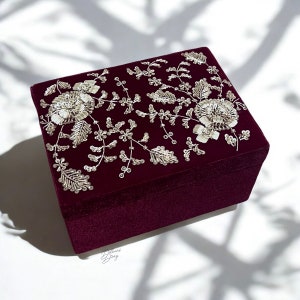 May include: A burgundy velvet jewellery box with a floral design embroidered in silver sequins. The box is closed and sitting on a white surface.